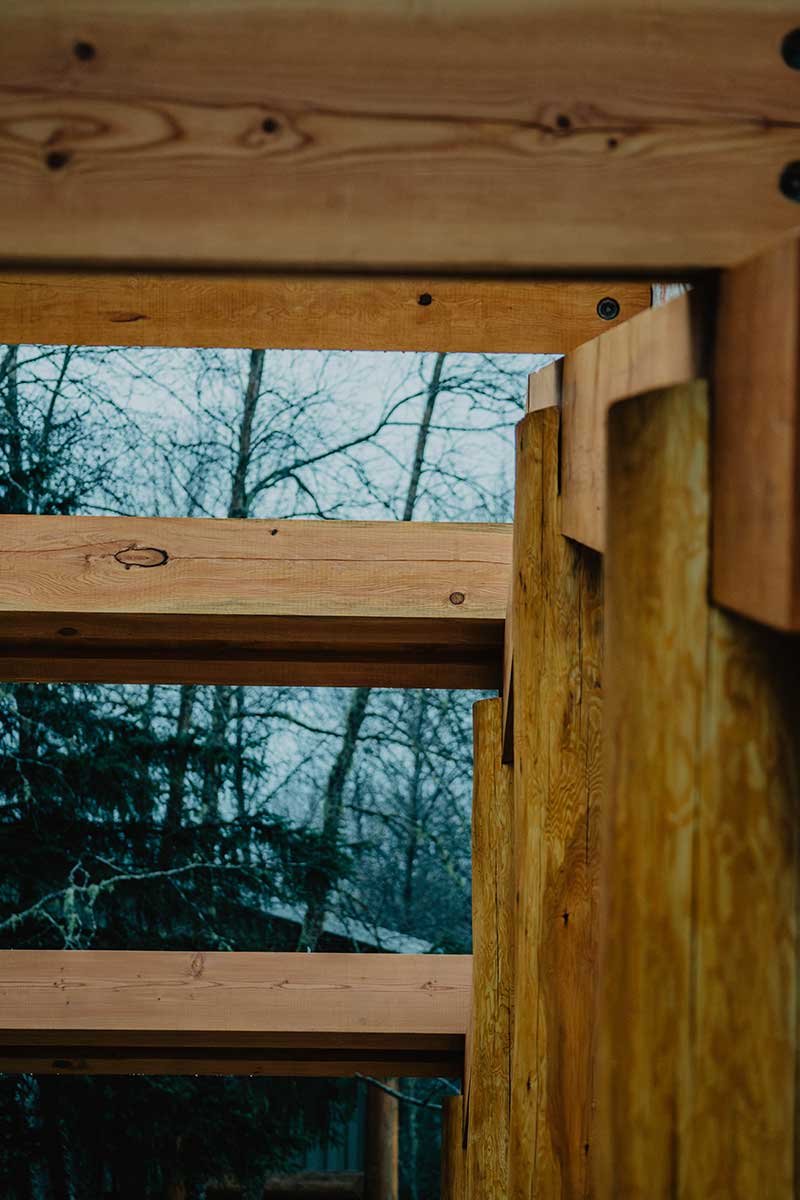 Timber frame structural elements with exposed wooden beams and joinery, viewed against a natural forest background, showcasing the raw craftsmanship of Smith Timber Works.