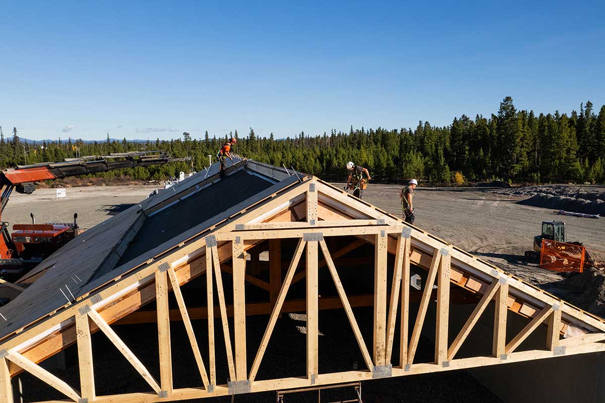 Aerial view of Smith Timber Works crew installing prefabricated framed trusses on a large prefabricated gable roof in a remote construction site.