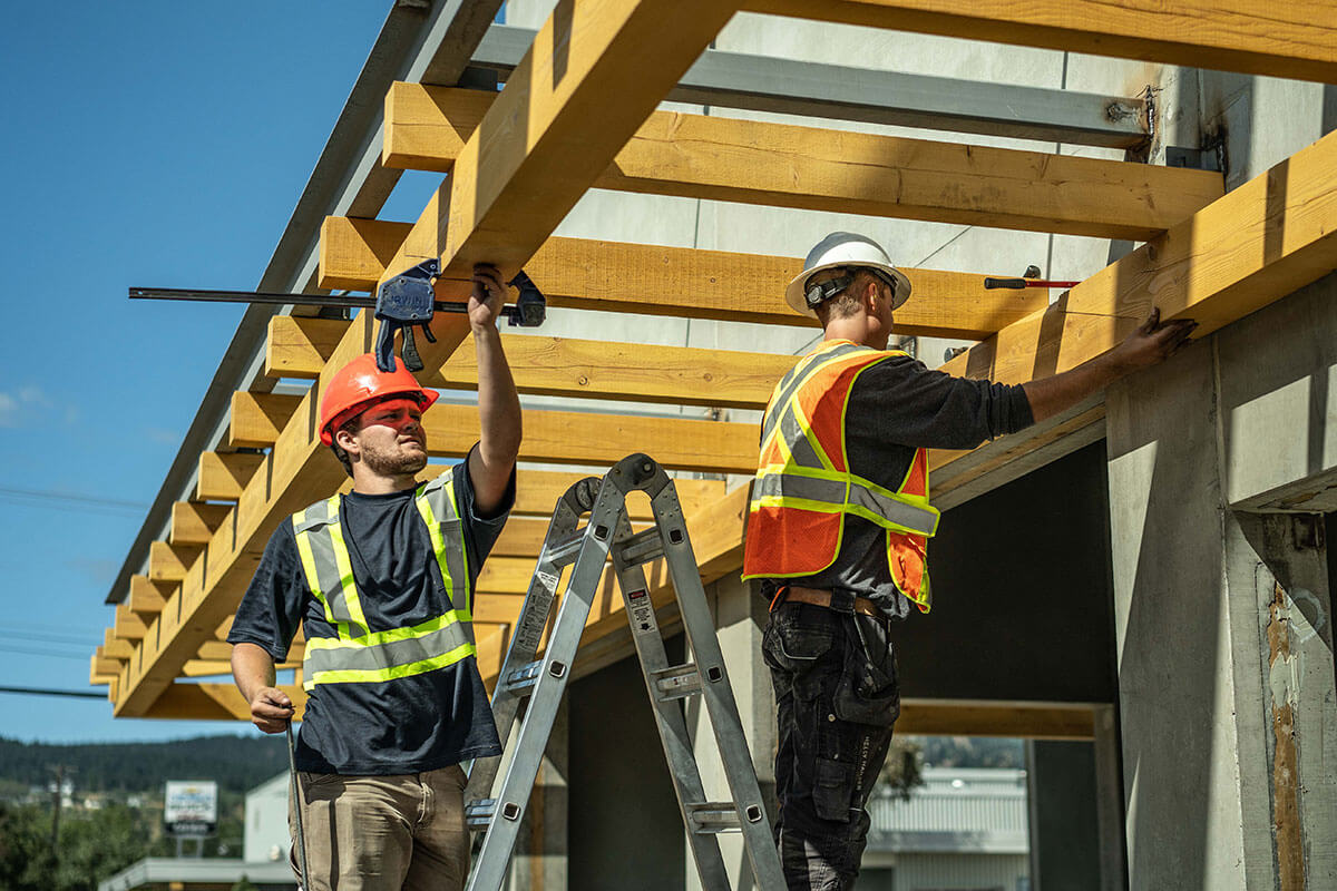 Two construction workers in safety vests and hard hats secure wooden beams on a commercial timber frame structure.