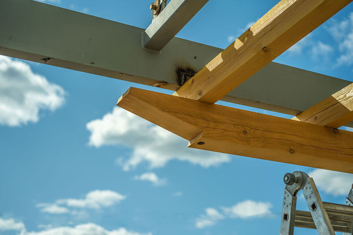 Close-up of a timber beam being installed onto a steel frame under a blue sky with clouds.