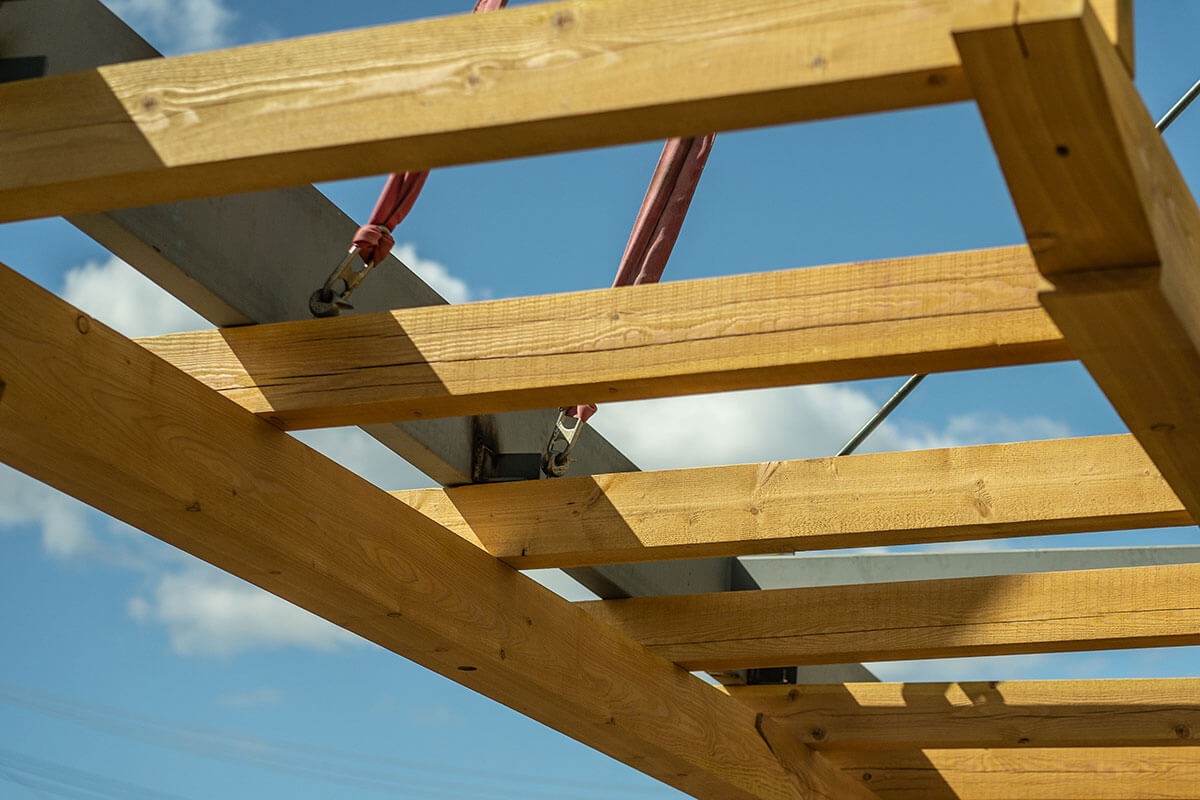 Close-up view of timber roof framing with steel beam and red lifting straps against a blue sky.