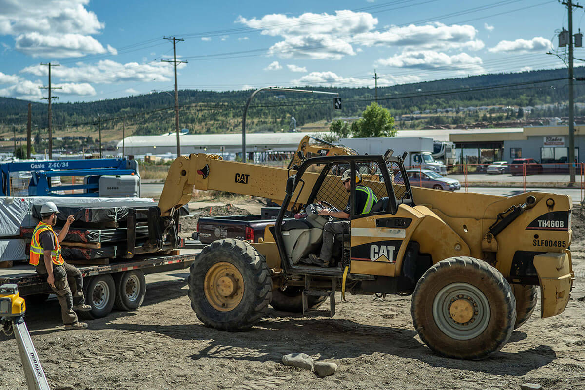 Construction site with a CAT telehandler unloading materials from a flatbed trailer while a worker supervises.