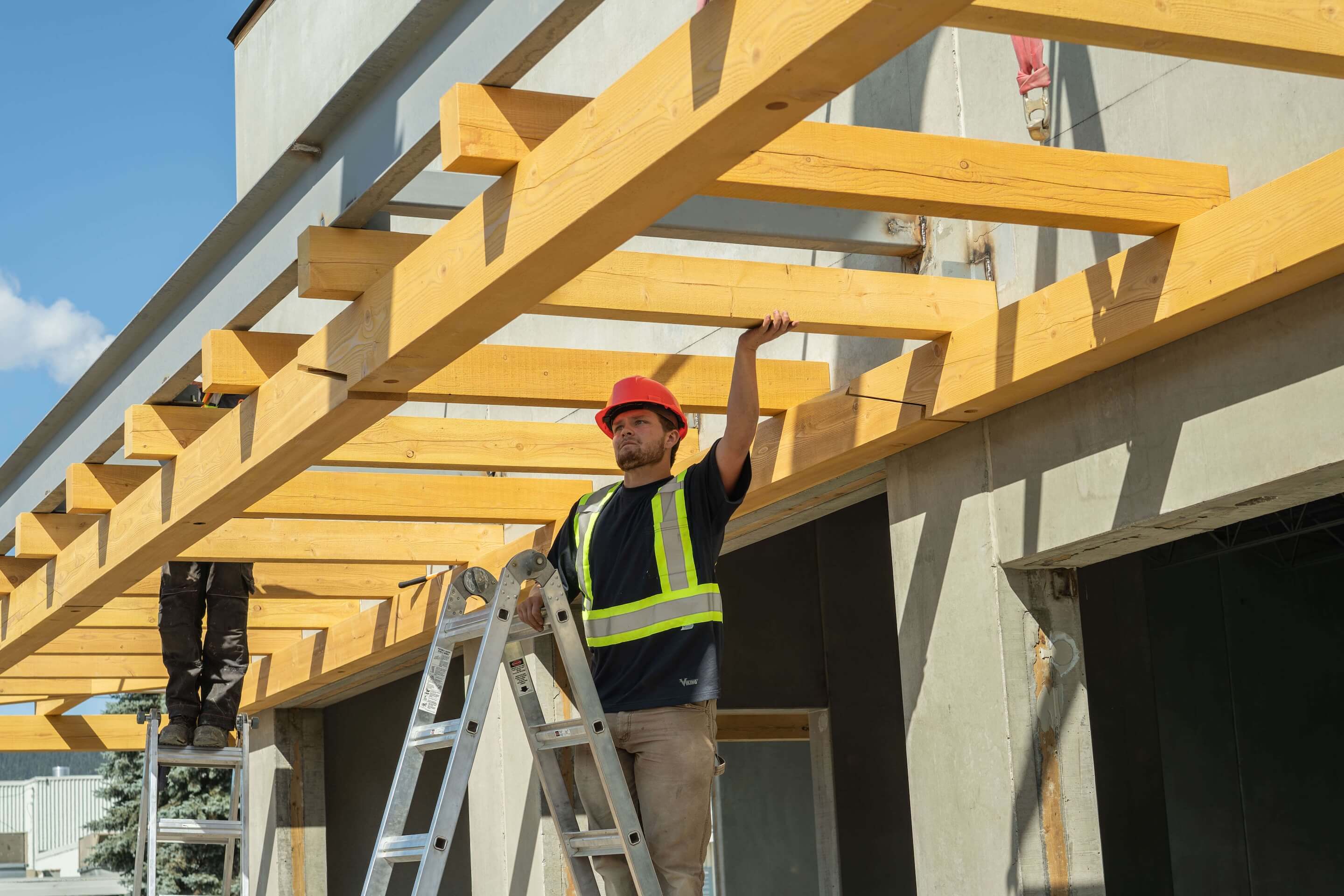 Construction worker in a red hard hat and safety vest aligning overhead timber beams on a commercial building site.