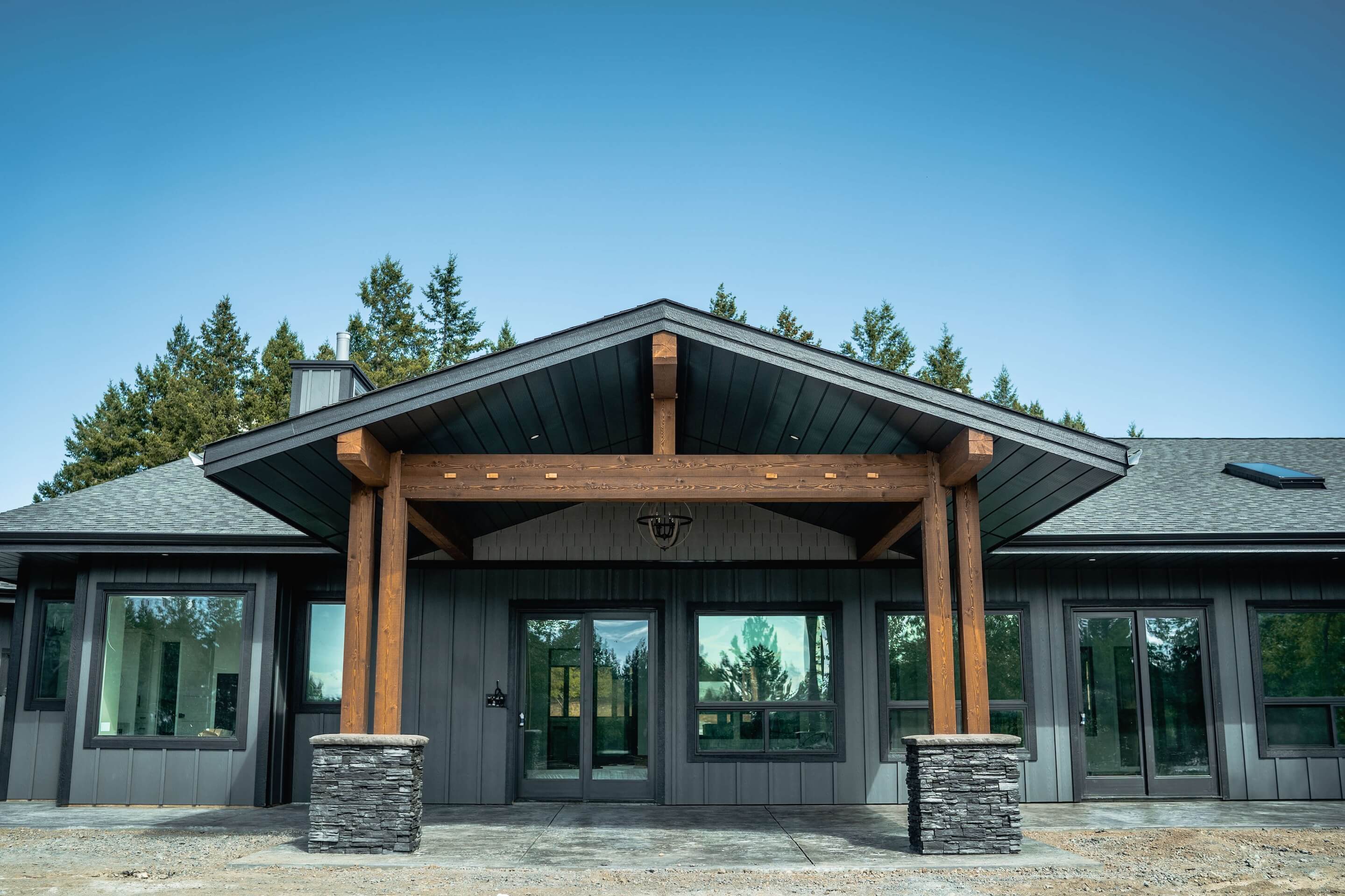 Modern dark-sided house with timber frame entry and stone column bases, set against a backdrop of pine trees and clear blue sky.
