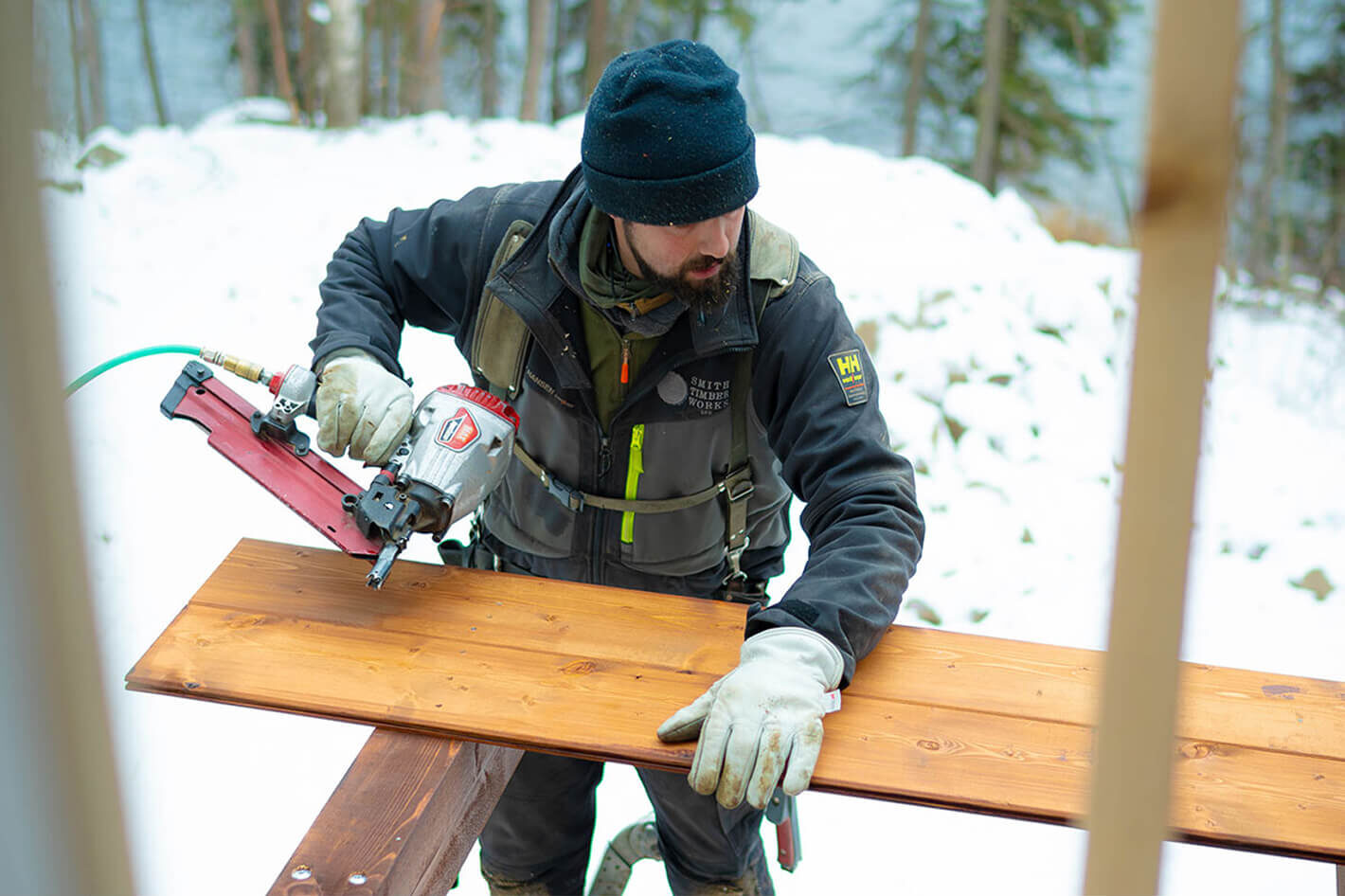 Timber framer fastening a stained beam with a nail gun during winter construction for Smith Timber Works in British Columbia