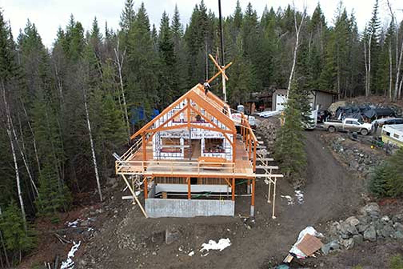 Aerial view of timber frame home under construction in a forested setting, with crane placement of structural beams by Smith Timber Works.