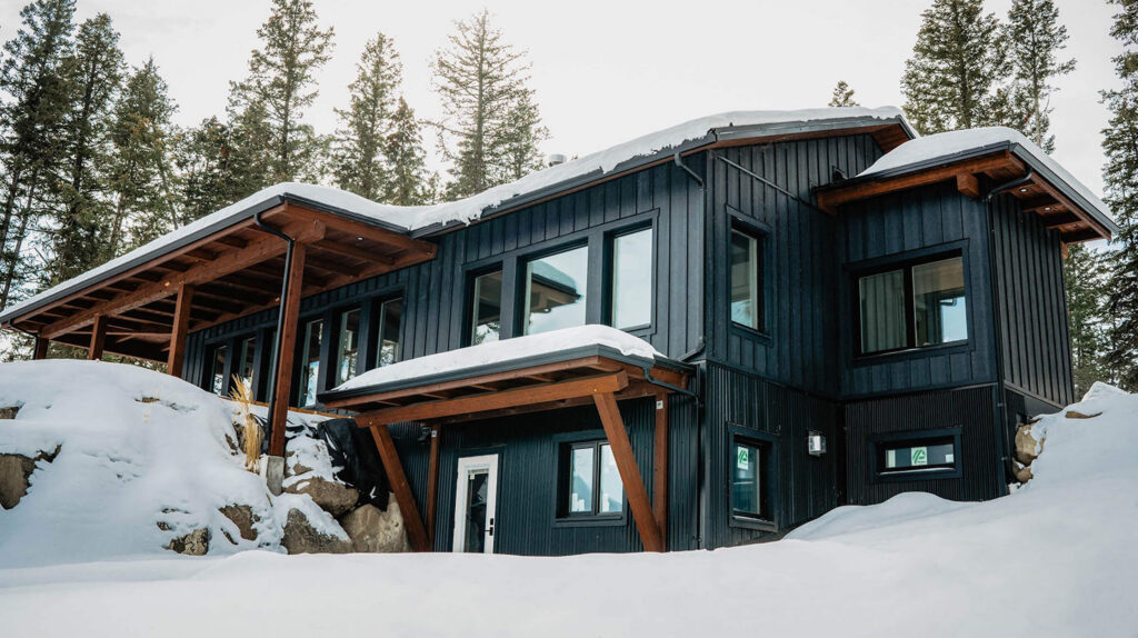 Snow-covered timber frame home with black siding and wood accents, built by Smith Timber Works in a forested landscape.