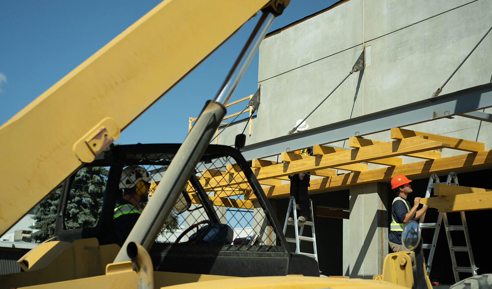 Construction workers installing yellow timber beams on a commercial building, with a telehandler in the foreground.