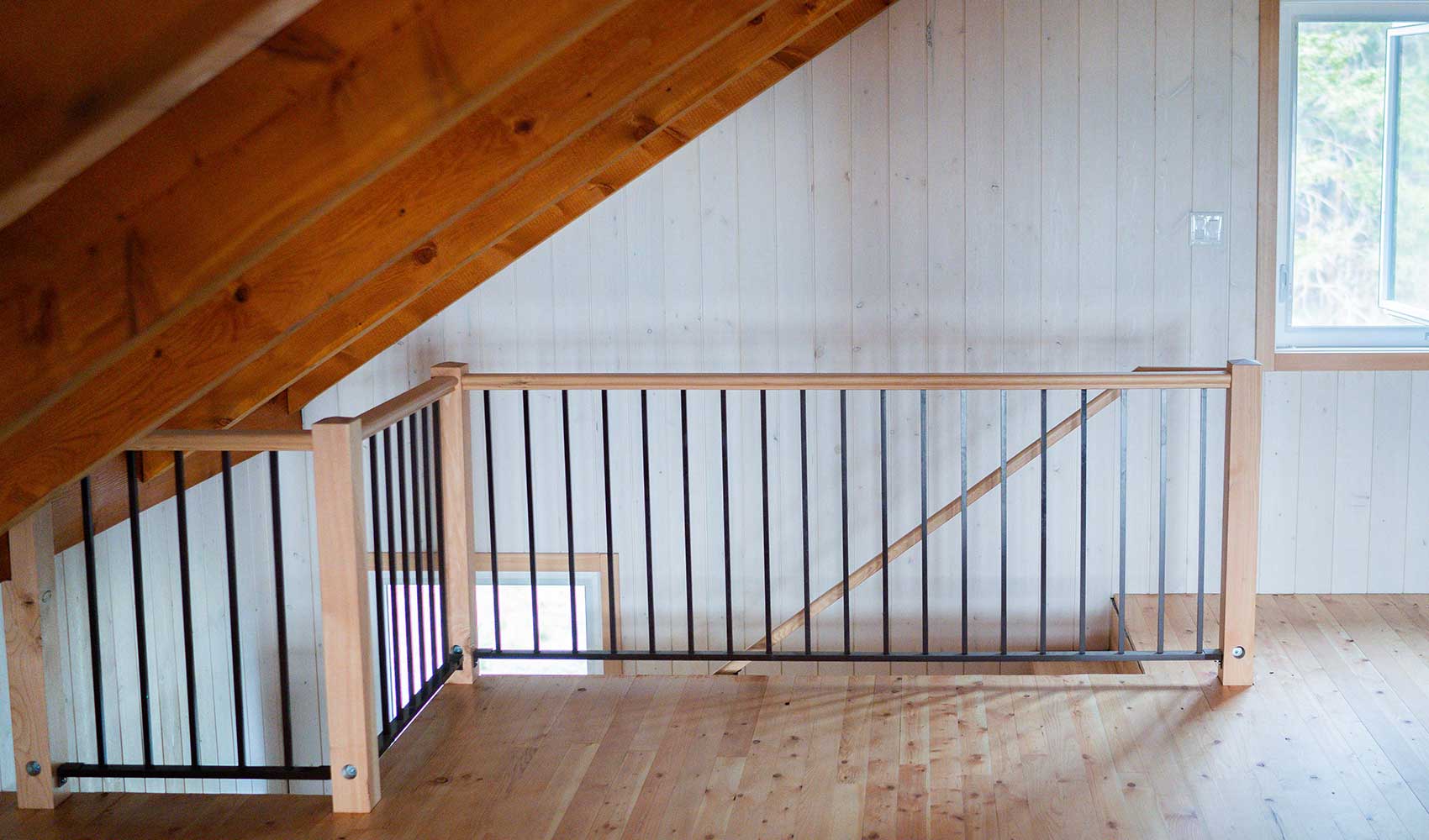 Loft area with sloped wooden ceiling, light wood floor, and black metal railing above a staircase.
