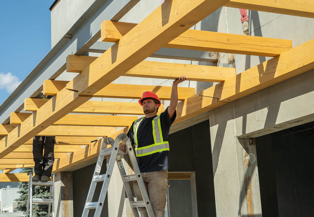 Construction worker in a red hard hat and safety vest aligning overhead timber beams on a commercial building site.