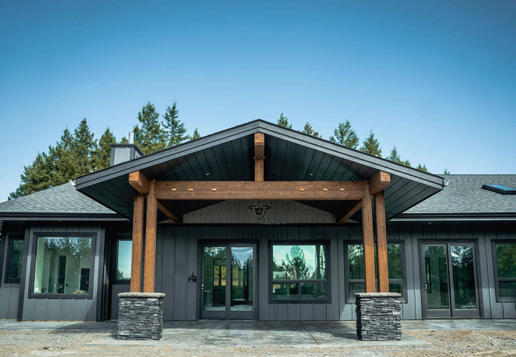 Modern dark-sided house with timber frame entry and stone column bases, set against a backdrop of pine trees and clear blue sky.