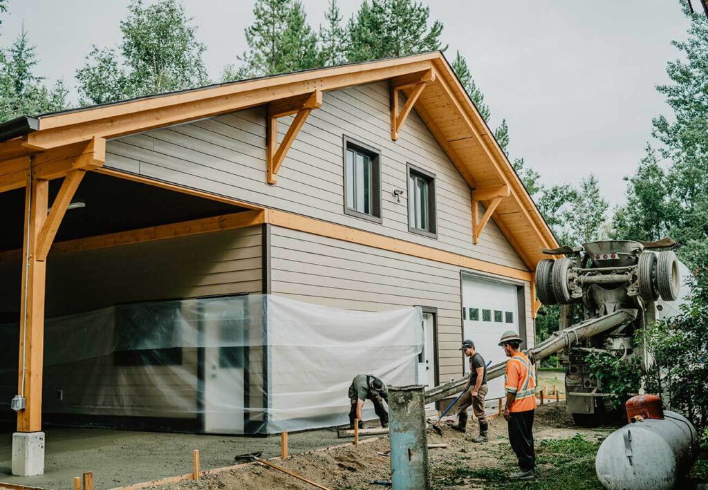 Canadian timber frame structure under construction by Smith Timber Works, featuring exposed wood accents, light siding, and on-site concrete work, highlighting design-build and prefabrication.